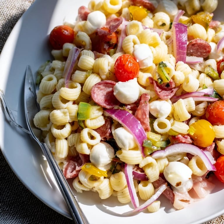 A close-up view of the Spring Antipasto Pasta Salad reveals chopped pickled vegetables, cherry tomatoes, and red onion mingling with pasta and greens.